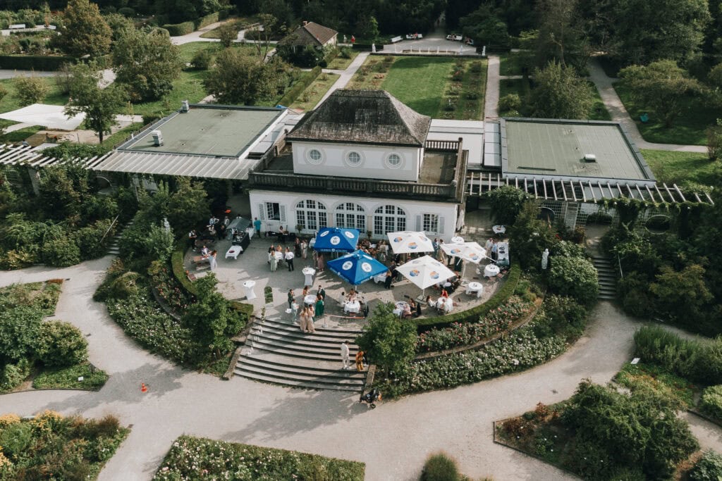 Hochzeitsgesellschaft im Parkrestaurant in München. Feierliche Atmosphäre mit Gästen unter Sonnenschirmen.