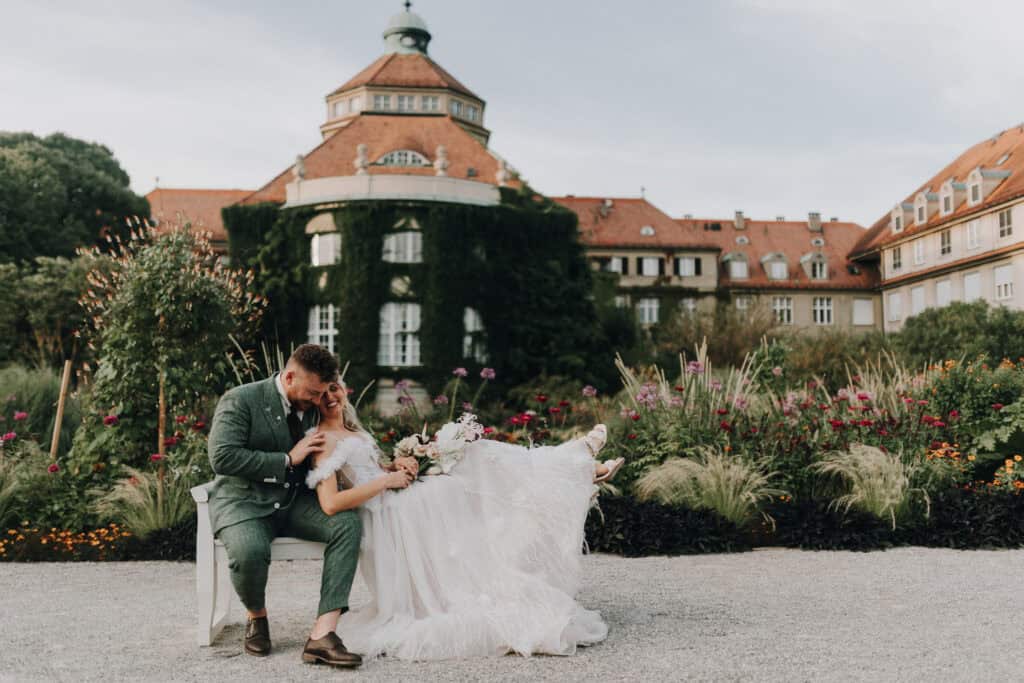 Brautpaar kuschelt auf Bank im Schlossgarten, München. Grün Anzug, helles Kleid. Glückliche Hochzeit.