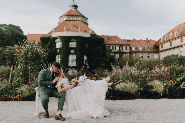 Brautpaar kuschelt auf Bank im Schlossgarten, München. Grün Anzug, helles Kleid. Glückliche Hochzeit.