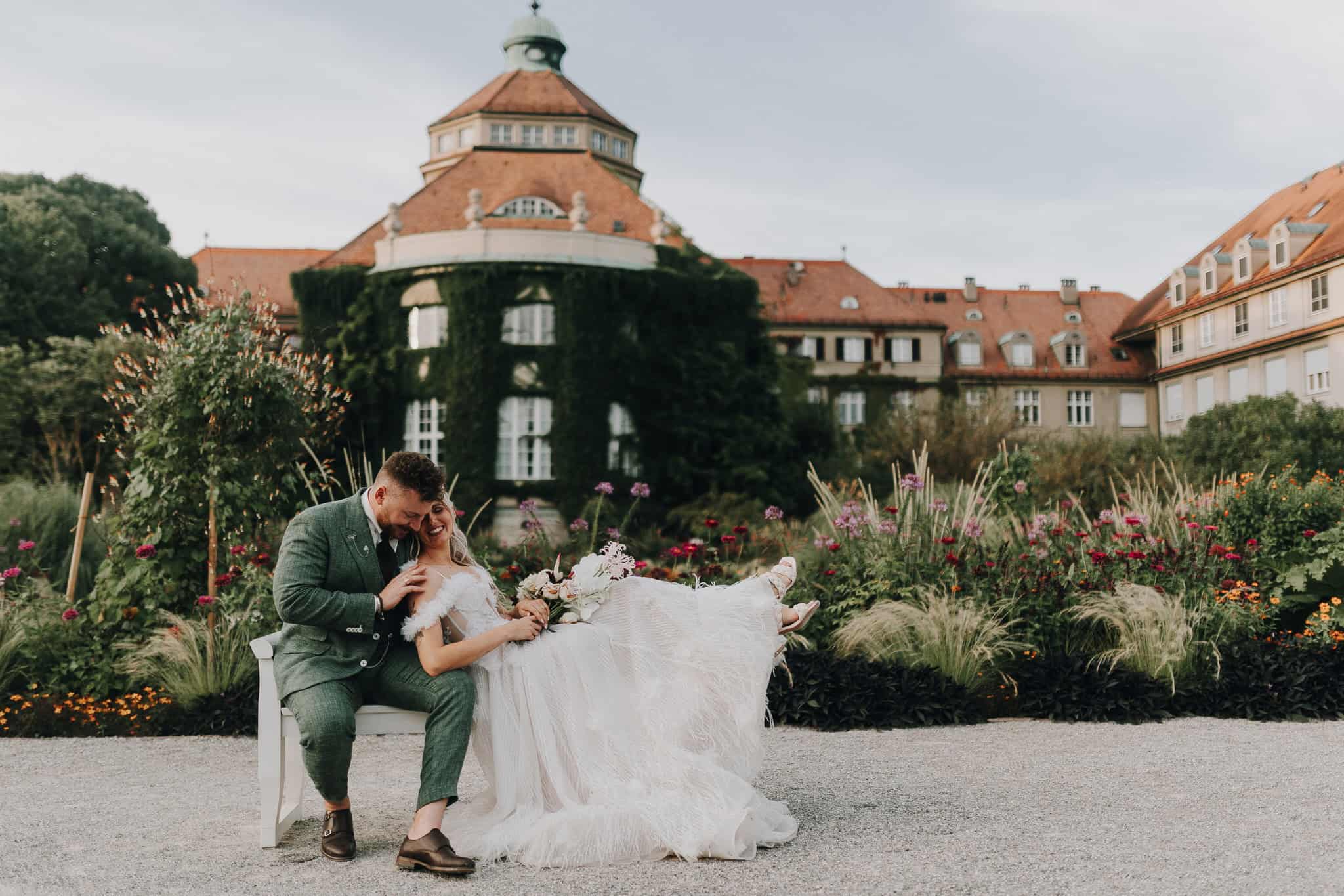 Brautpaar kuschelt auf Bank im Schlossgarten, München. Grün Anzug, helles Kleid. Glückliche Hochzeit.