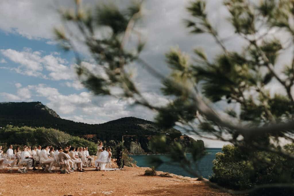 Hochzeitszeremonie im Freien auf Ibiza mit Blick auf das Meer und die Berge. Gäste sitzen auf Stühlen.