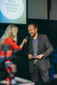 Mann nimmt Auszeichnung entgegen, Moderatorin mit Mikrofon. Preisverleihung, beste Fotografie Brautpaar-Portrait.