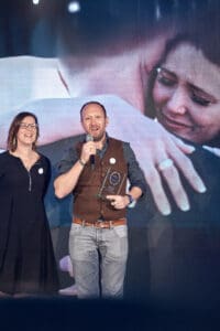 Mann nimmt Preis entgegen, Frau steht daneben. Paar im Hintergrund auf Leinwand. Preisverleihung.