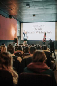 Präsentation vor Publikum mit dem Titel "Die Basics: G wie Gerade Wirbelsäule" auf einer Leinwand. Hochzeitsfotografie Workshop