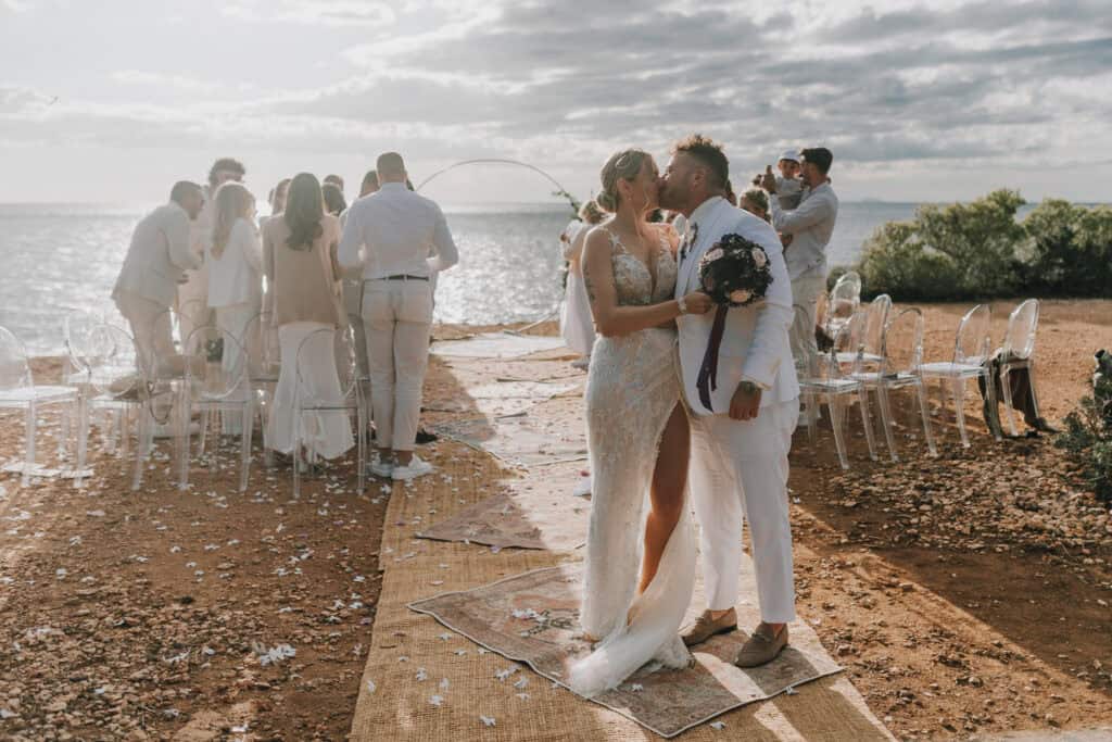 Brautpaar küsst sich bei Strandhochzeit auf Ibiza. Gäste in Weiß. Zeremonie am Meer. Romantisch!