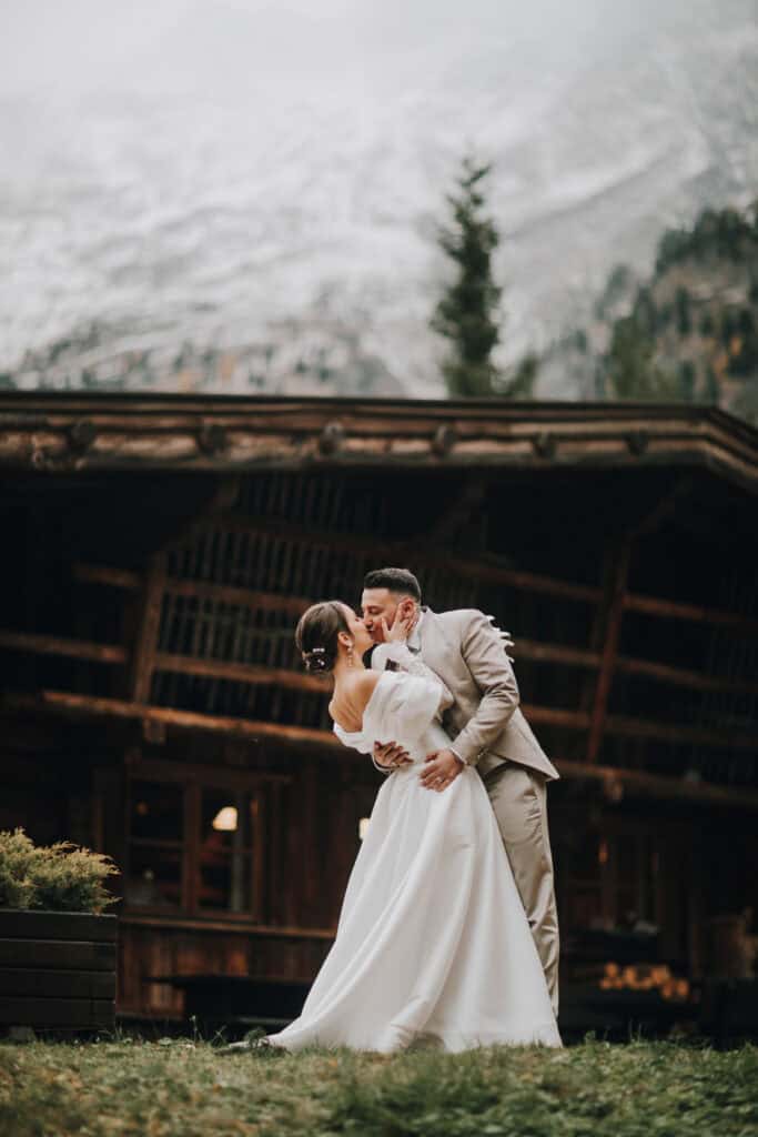 Brautpaar küsst sich vor rustikalem Holzhaus in Tirol. Romantische Hochzeit in den Bergen.