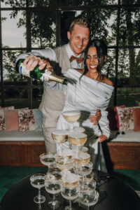 Brautpaar gießt Sekt in eine Sektpyramide, lachend. Hochzeitsfotograf Tirol könnte hier sein.