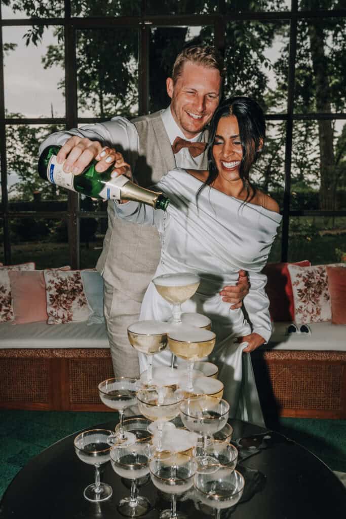 Brautpaar gießt Sekt in eine Sektpyramide, lachend. Hochzeitsfotograf Tirol könnte hier sein.