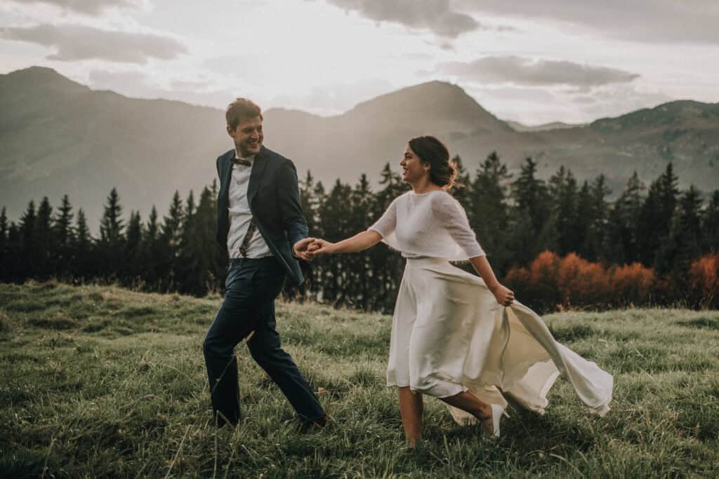 Lächelndes Brautpaar hält Händchen und rennt über eine Wiese mit Bergen im Hintergrund. Hochzeitsfotograf Tirol.