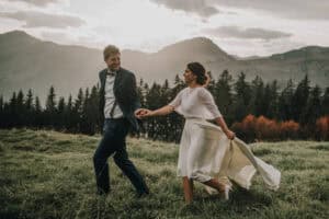 Lächelndes Brautpaar hält Händchen und rennt über eine Wiese mit Bergen im Hintergrund. Hochzeitsfotograf Tirol.