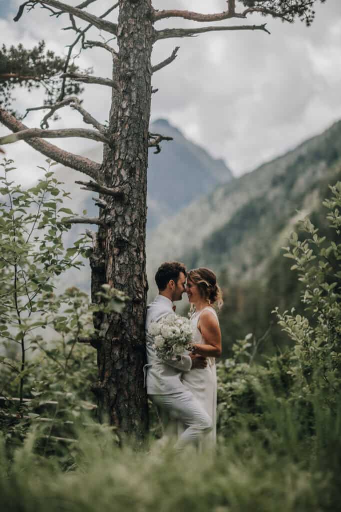 Brautpaar umarmt sich vor Baum in Tiroler Berglandschaft mit weißem Brautstrauß.