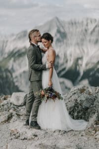 Hochzeitspaar umarmt sich in den Bergen Tirols. Braut mit Brautstrauß, Bräutigam küsst sie auf die Stirn.