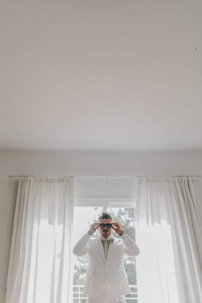 Bräutigam im weißen Anzug setzt Sonnenbrille vor Fenster auf. Hochzeitsfotograf Tirol.