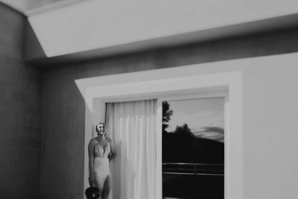 Lachende Braut im Brautkleid vor Fenster. Hochzeitsfotograf Tirol.