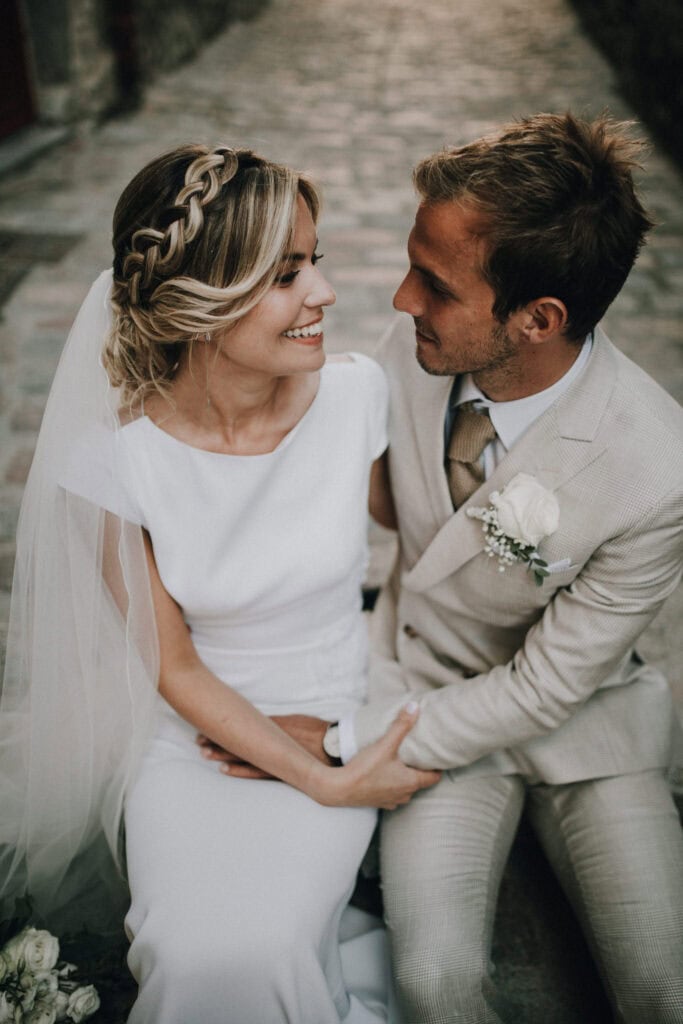 Brautpaar sitzt eng umschlungen, lächelt sich an. Brautkleid, Anzug, Schleier. Hochzeitsfotograf Tirol.