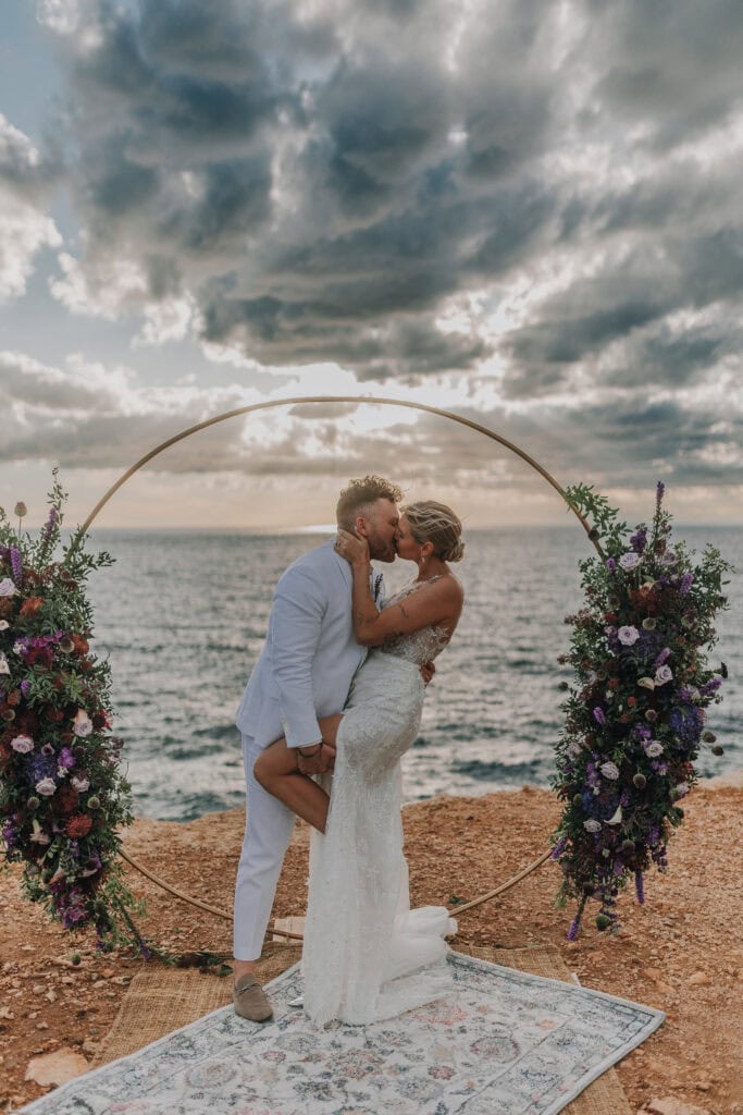 Brautpaar küsst sich vor Meer und Blumenbogen. Hochzeitsfotograf Tirol.