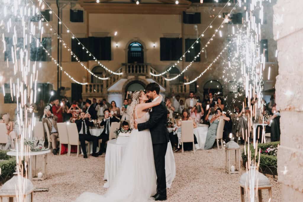 Brautpaar küsst sich vor Hochzeitstorte und Publikum, umgeben von Fontänen.