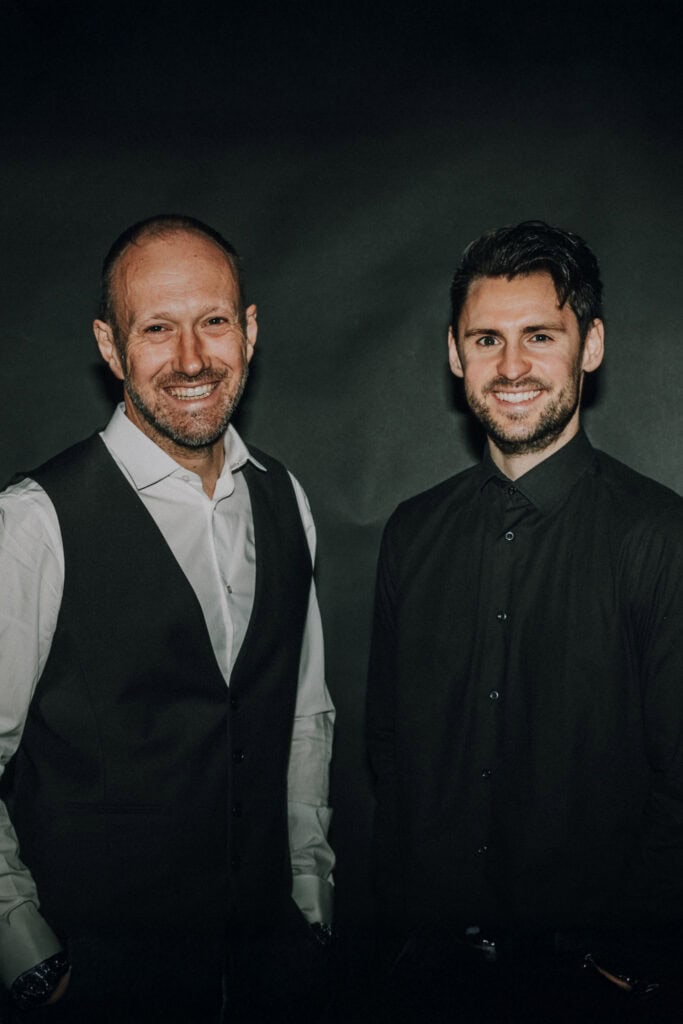 Zwei Männer in formeller Kleidung lächeln vor dunklem Hintergrund. Hochzeitsfotograf Tirol könnte relevant sein.