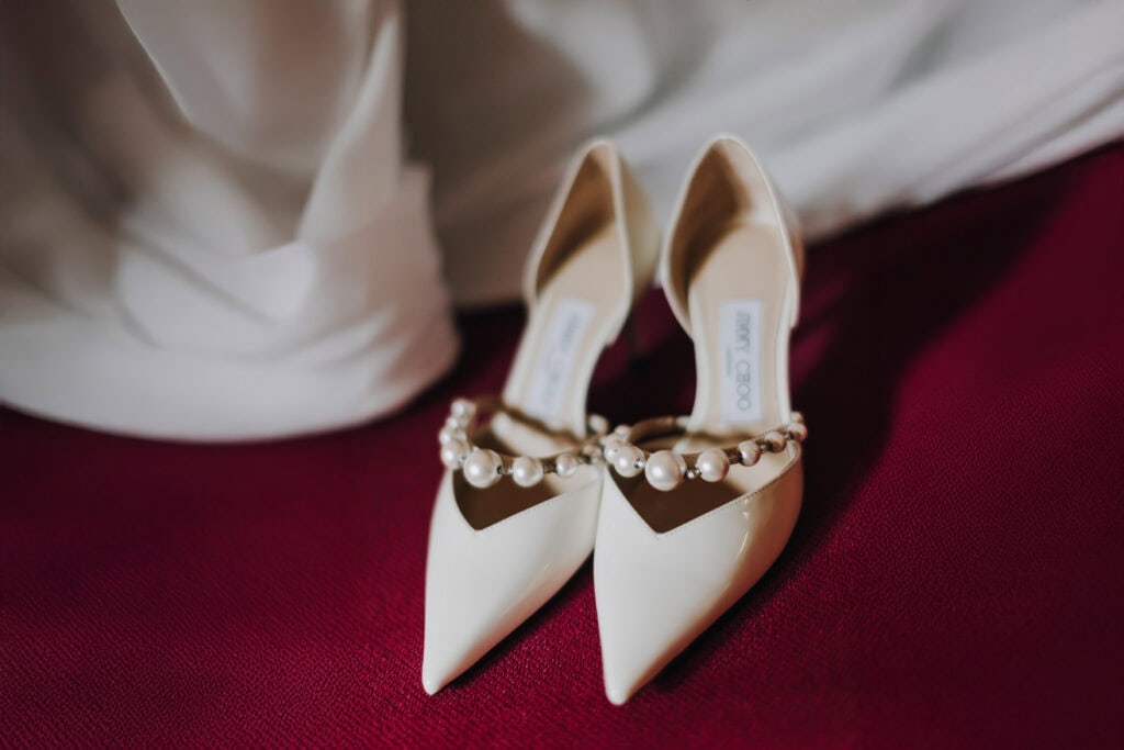 Elegante elfenbeinfarbene Jimmy Choo Brautschuhe mit Perlen auf bordeauxfarbenem Hintergrund.